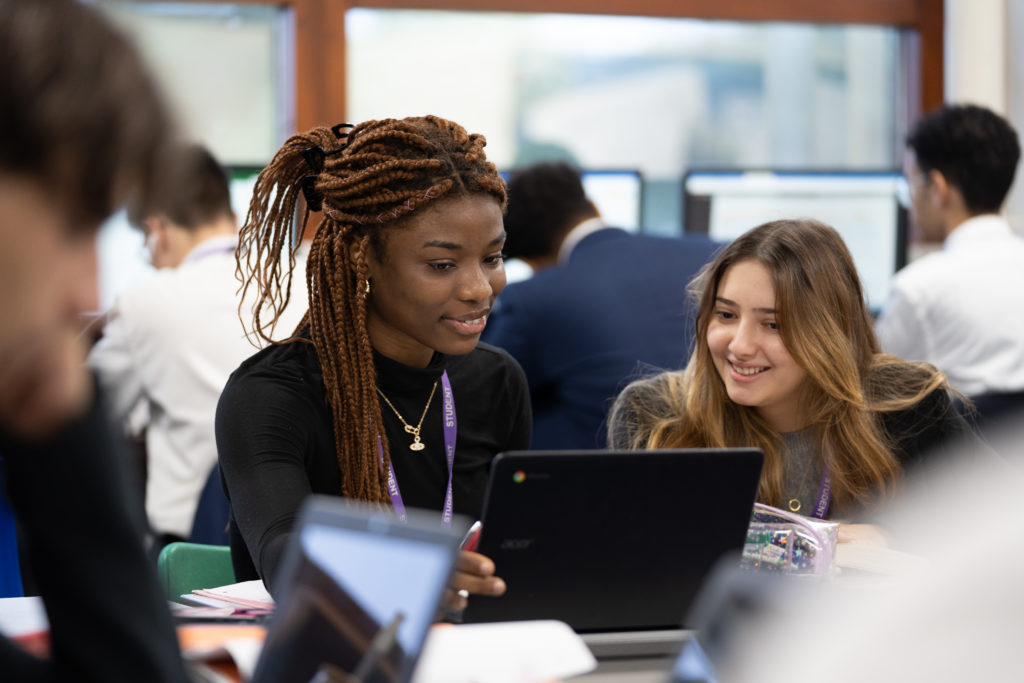 Two girls are seen sitting next to one another at a desk, smiling and looking at a laptop they are sharing.