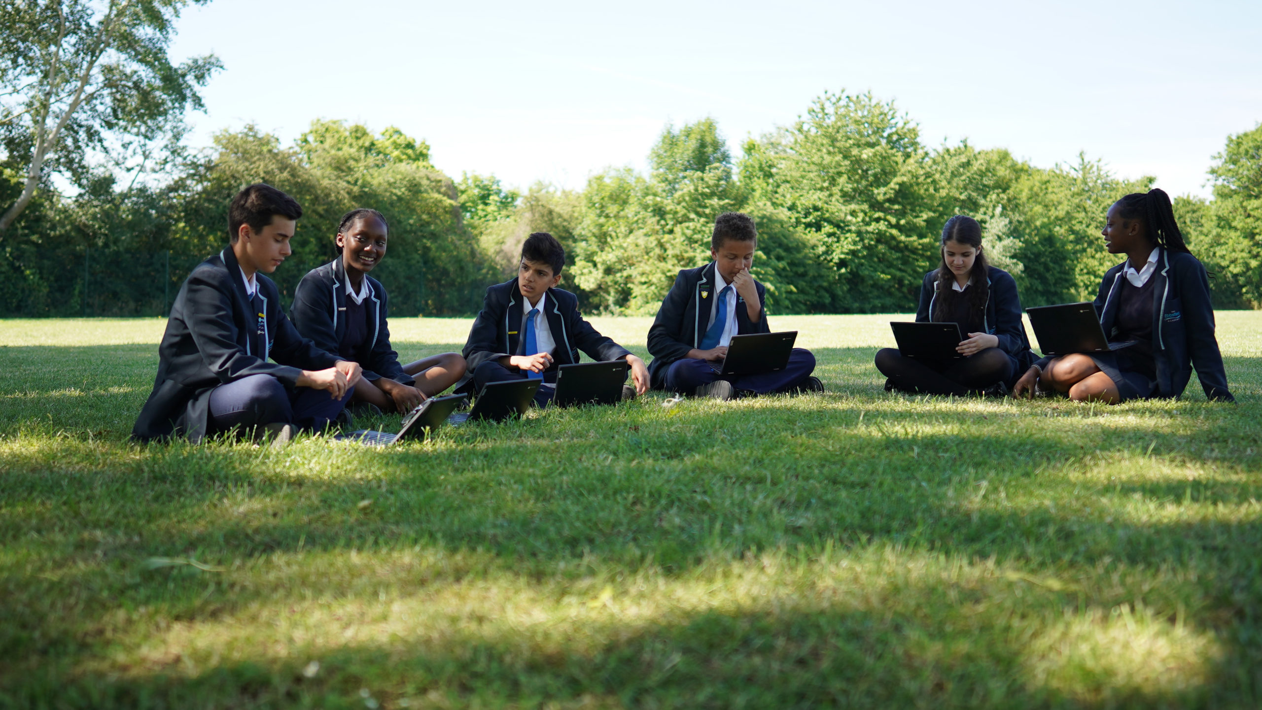Six Leigh Academy Ebbsfleet students are seen sitting on a grass area outside and using their laptop computers.