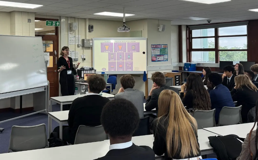 Here's a breakdown of what's in the picture: Teacher: On the left side of the room, a woman with dark hair and glasses is standing in front of a whiteboard, gesturing with her hands as she speaks. She appears to be leading a lesson. She is wearing dark clothing and has an ID badge. Students: A group of students, mostly teenagers, are seated at desks arranged in rows, facing the front of the classroom. Many of them are looking towards the teacher and the front of the room. Some are wearing school uniforms (dark blazers). Classroom Environment: Whiteboards: There's a large whiteboard on the left, partially filled with writing, and another large interactive whiteboard or screen at the front of the room displaying a presentation with illustrations (possibly related to "units" or measurements). Projector: A projector is mounted on the ceiling, pointed towards the main screen. Desks and Chairs: Individual desks and chairs are arranged for the students. Windows: Windows are visible on the right side of the room, letting in natural light. Posters/Decorations: Various posters and charts are on the walls, typical of a classroom. Door: A door with an "Exit" sign above it is visible in the background on the left. Supplies: Some school supplies or boxes are visible near the windows.