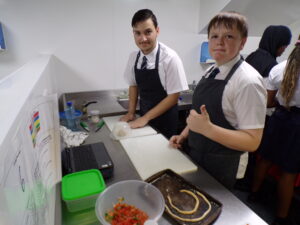 Two male students are at a counter. One is kneading or shaping dough under a clear bowl, while the other gives a thumbs-up gesture to the camera. There's a tray with a piece of shaped dough, and a bowl of chopped tomatoes is in the foreground, indicating pizza or a similar dish being prepared.