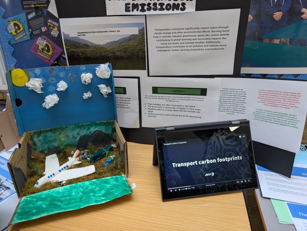 This image focuses on a project related to transportation emissions and carbon footprints. It includes: A poster titled "TRANSPORTATION EMISSIONS IMPACT ON NATURE." A physical diorama or model featuring a small airplane, possibly illustrating a landscape impacted by emissions. A laptop screen displaying a video titled "Transport carbon footprints." Additional text on posters discussing community impacts and effects of carbon emissions.