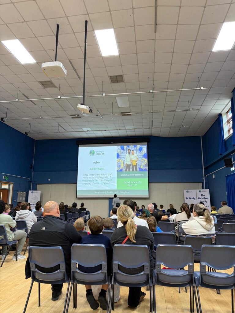 Picture shows a large group of people, including what look like parents and children, are seated in a hall, facing a stage with a large screen. The screen in the first image shows a student's name, "Ayham," and a quote about their academic success. The photo seems to capture a formal school event.