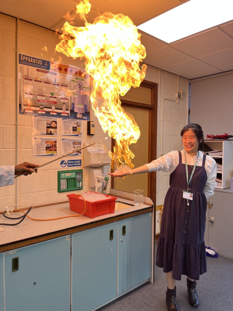 picture is a classroom or laboratory setting where a student is participating in a science experiment. A large flame is rising from a small, bubbly-looking pile in her hand. Another person, whose hand and sleeve are visible, is holding a long rod, possibly a tool to ignite the substance. This image seems to show a science demonstration, likely a fun and exciting one involving fire.