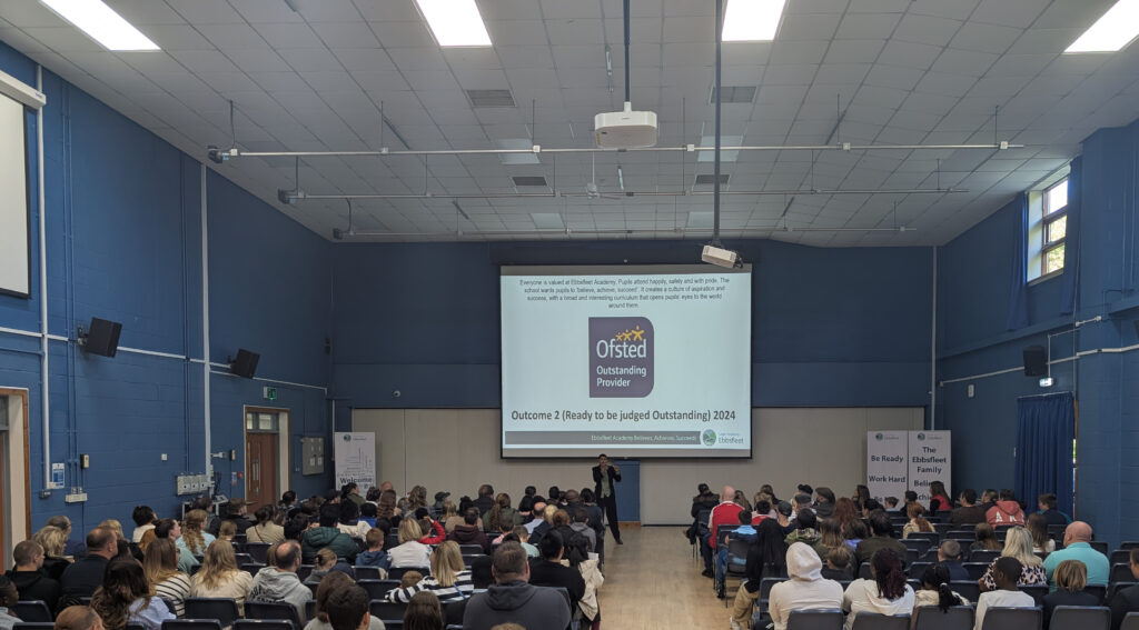 Picture shows a large group of people, including what look like parents and children, are seated in a hall, facing a stage with a large screen. The image has the text "Ofsted Outstanding Provider" and a quote about the school. The photo seems to capture a formal school event, possibly an awards ceremony or a presentation.