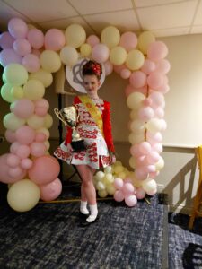 Young girl posing in front of a balloon arch with 1st place trophy wearing red, white and gold Irish dancing dress.