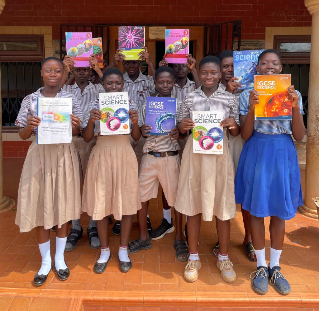 A group of students, mostly in striped uniforms, stand outside a building, holding various colorful textbooks, primarily science books. Some students hold books in front of their chests while others hold them above their heads.