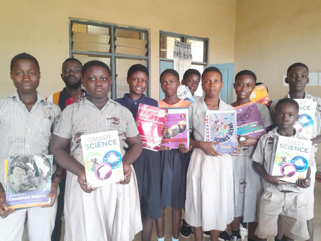 A group of students, mostly in striped uniforms, stand outside a building, holding various colorful textbooks, primarily science books. Some students hold books in front of their chests while others hold them above their heads.