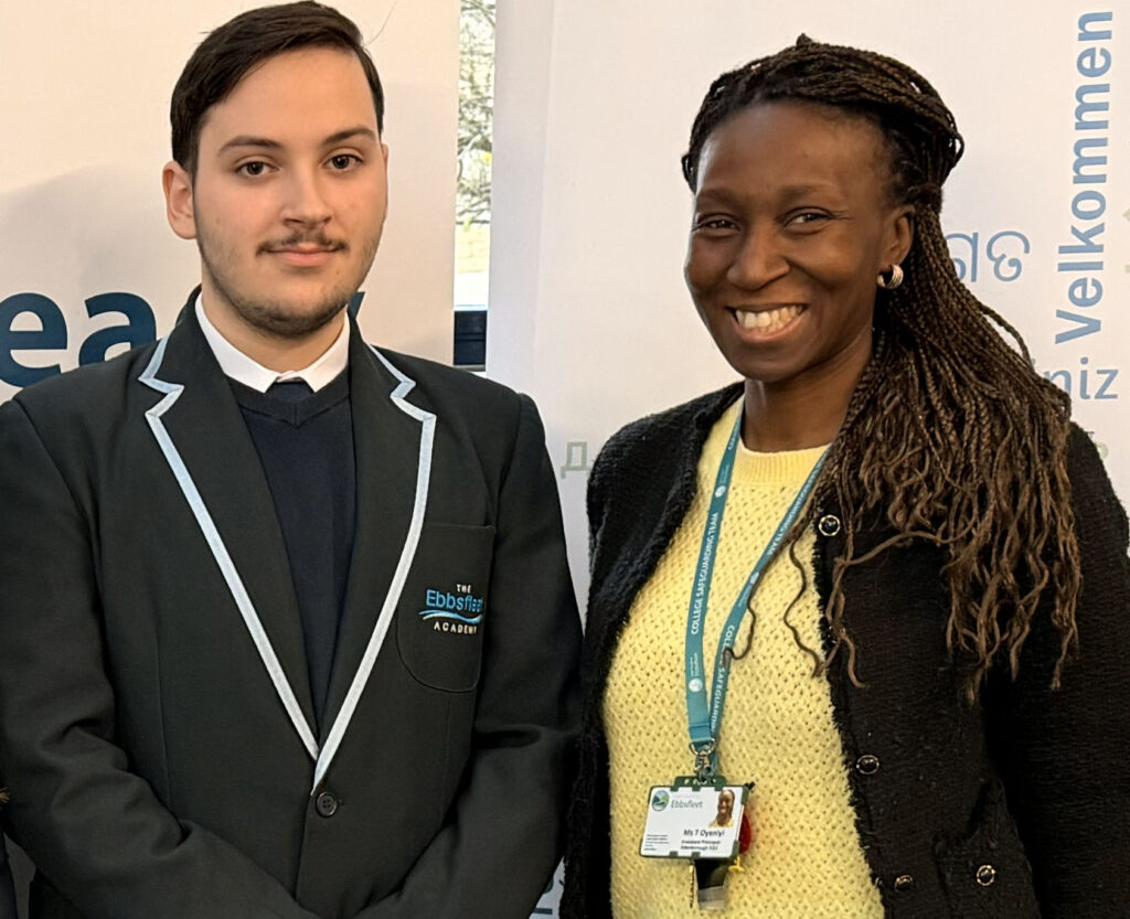 A young man in a black and blue school blazer from "The Ebbsfleet Academy" standing next to a smiling woman with braided hair, who is wearing a yellow sweater, a black cardigan, and a lanyard with an ID badge that reads "Ms T Oyeniyi"