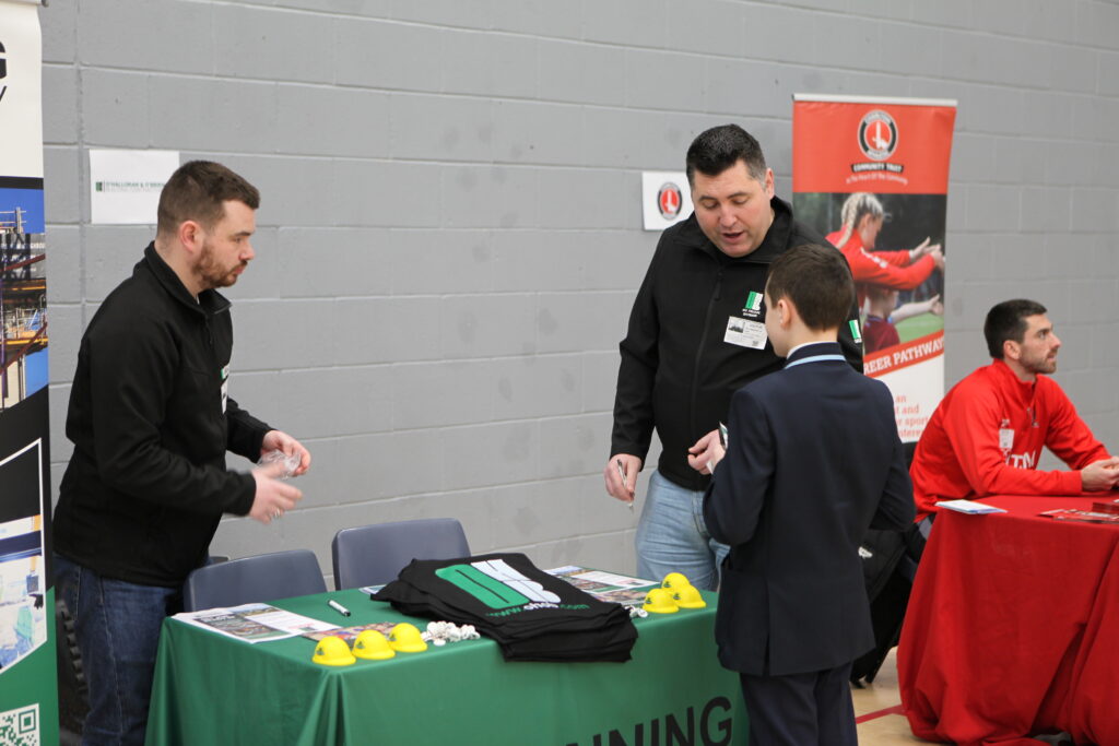 Two men at a construction company booth talk to a student; the table features miniature yellow hard hats and branded apparel.