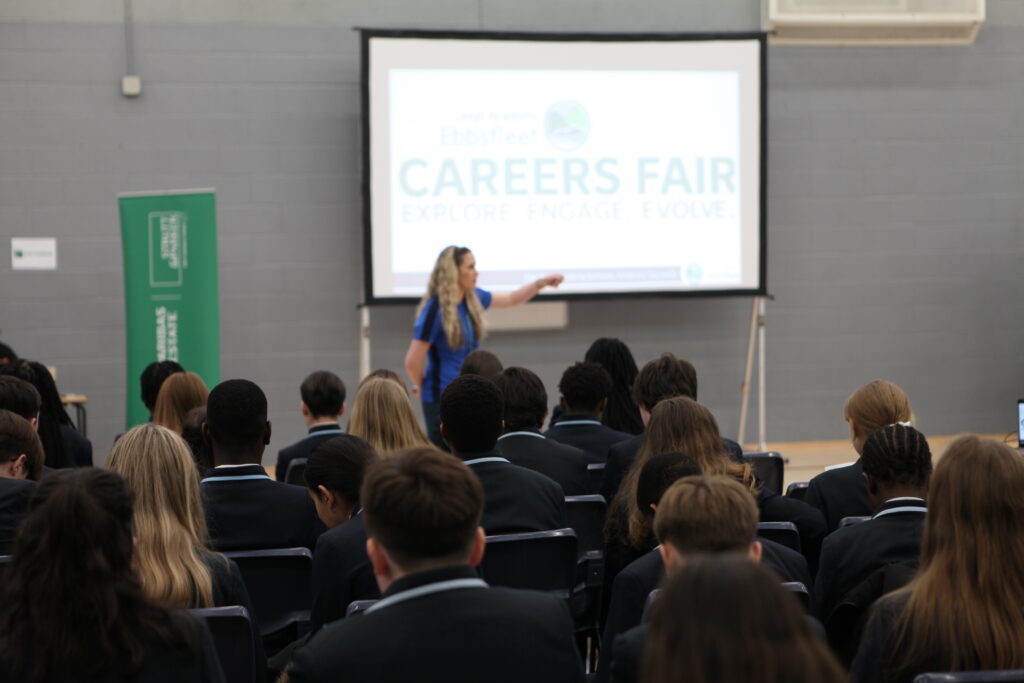 A woman presents to a large seated audience of students in uniform; a projector screen behind her displays "Careers Fair: Explore, Engage, Evolve."
