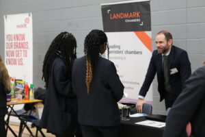 Two students with braided hair speak with a male professional at a career fair stand for Landmark Chambers.