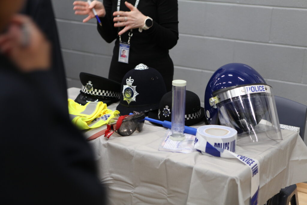 A display table at a career fair featuring various police equipment, including helmets, high-visibility gear, and a riot shield.