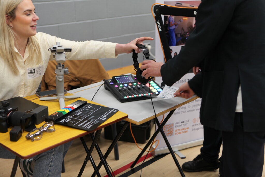 A female professional at a media booth hands a pair of studio headphones to a student over a table with a sound mixer and camera equipment.