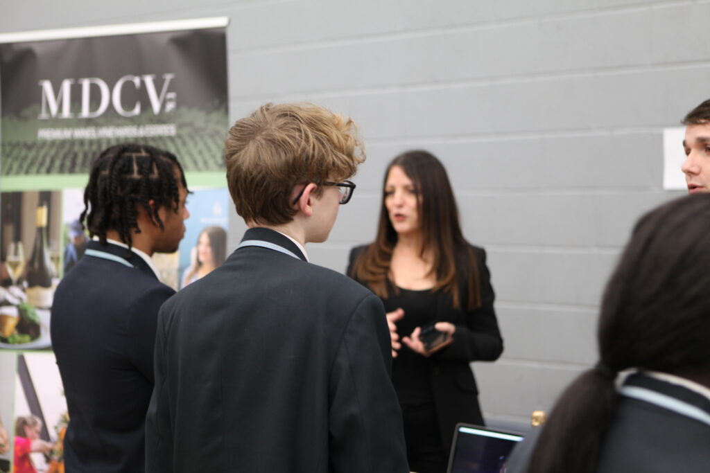 A female representative speaks with two students in school blazers at a career fair booth for a wine and estates company.