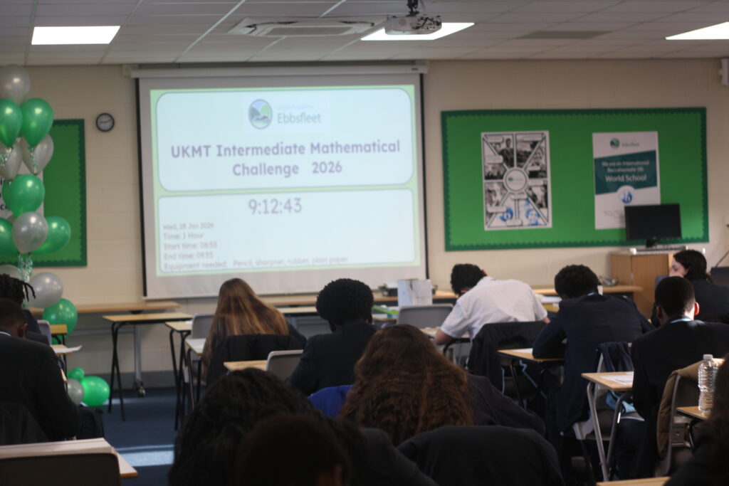 A wide classroom view of students working at their desks during a math competition, with a projector screen showing the event title and a digital timer.