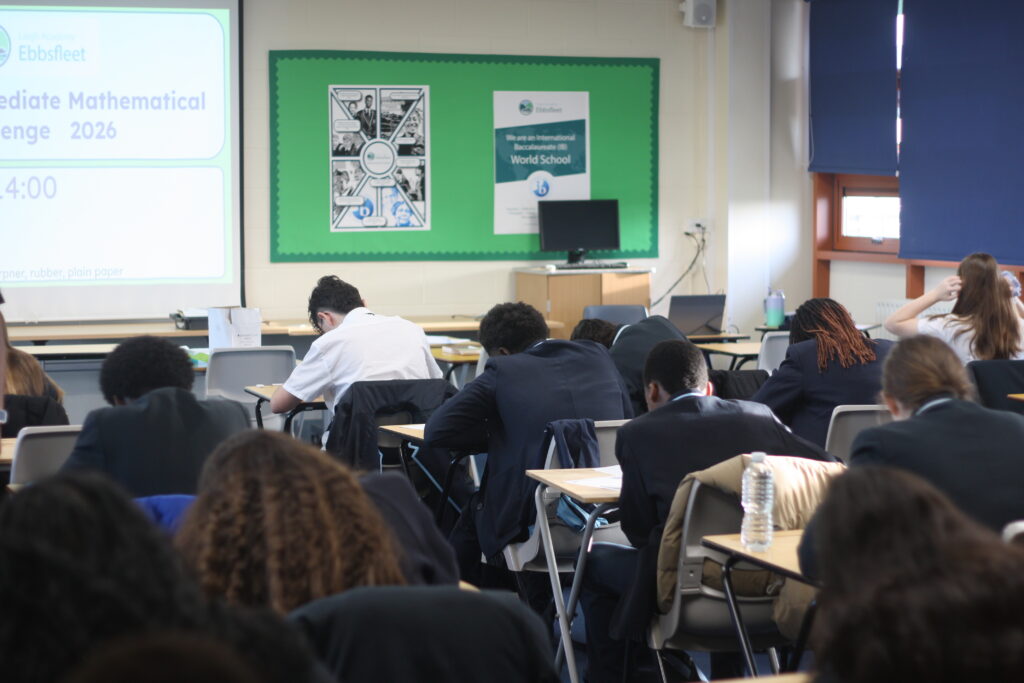 A focused shot of students in school uniforms working on a test at their desks, with a green notice board and a projector screen in the background.