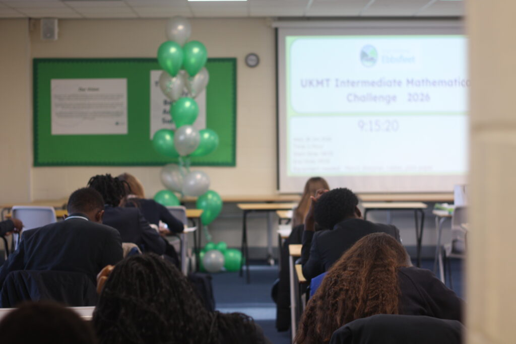 Students sitting at desks in a classroom, facing a projector screen that displays "UKMT Intermediate Mathematical Challenge 2026" next to a bunch of green and silver balloons.