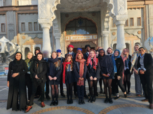 A wide-angle, eye-level outdoor shot shows a group of about 20 people, most of them teenagers, standing in front of a large, ornate building with white pillars and intricate carvings. The building has a large arched entrance with a gold-colored sign above it that has writing on it. The group is arranged in two rows, with the taller people in the back and the shorter people in the front. Most of the people are wearing dark-colored clothing, and many of the girls are wearing headscarves of various colors, including black, green, blue, pink, and red. Some of the boys are wearing turbans. The group is smiling and looking at the camera. The ground is paved with light-colored stones, and there is a large, metallic sculpture to the left of the group. The sun is shining, and there are long shadows on the ground. The sky is clear and blue. The overall mood of the image is positive and inclusive.