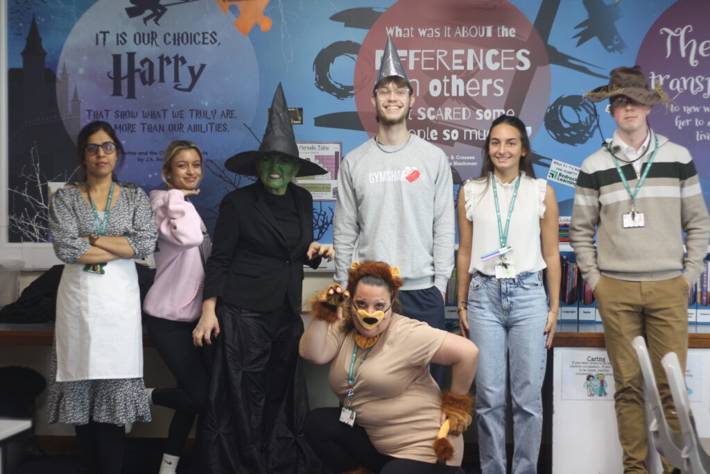 A group of seven adults in various costumes, including a witch and a scarecrow, posing in front of a Harry Potter-themed wall display.