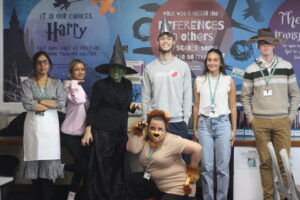 A group of seven adults in various costumes, including a witch and a scarecrow, posing in front of a Harry Potter-themed wall display.