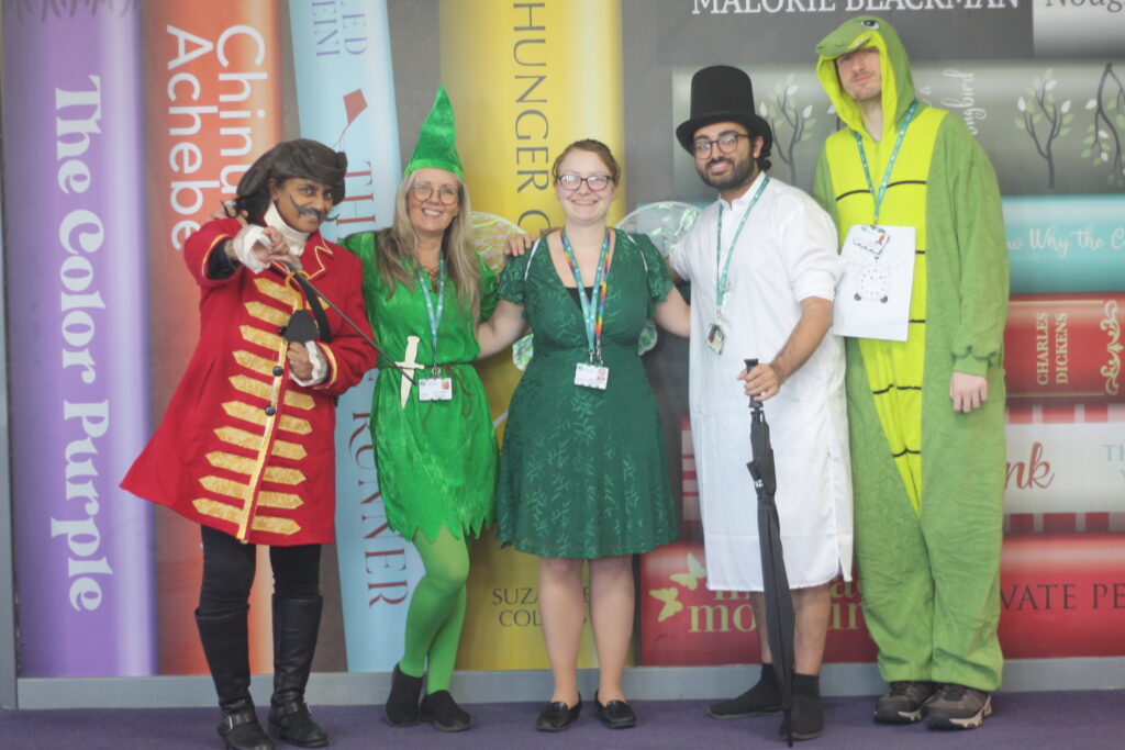 Five adults dressed in costumes, such as a pirate, a fairy, and a crocodile, standing in front of a wall painted to look like giant book spines.