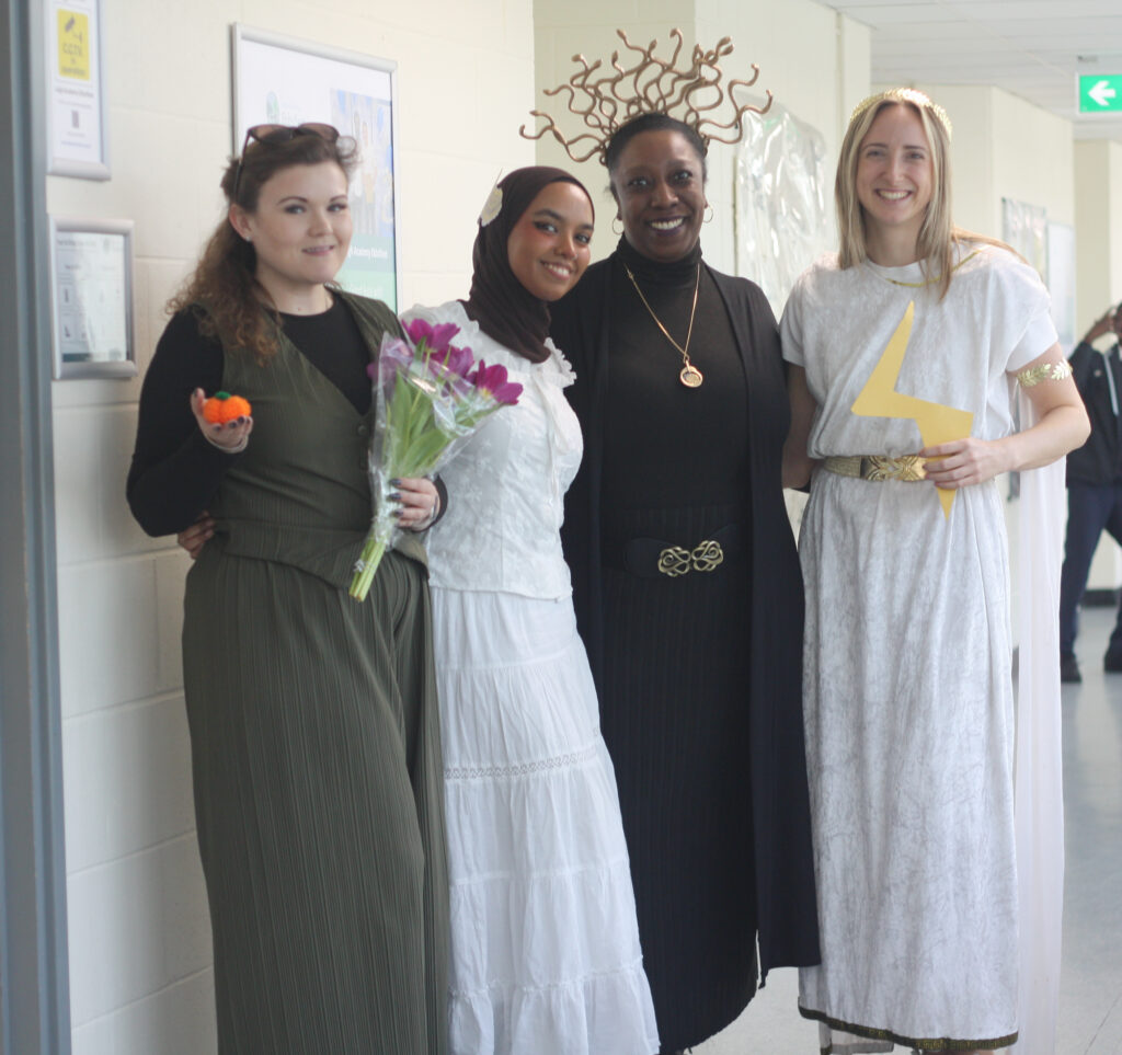 Four adults posing in a hallway in different outfits, including one person with a snake headpiece and another in a white toga holding a yellow lightning bolt.