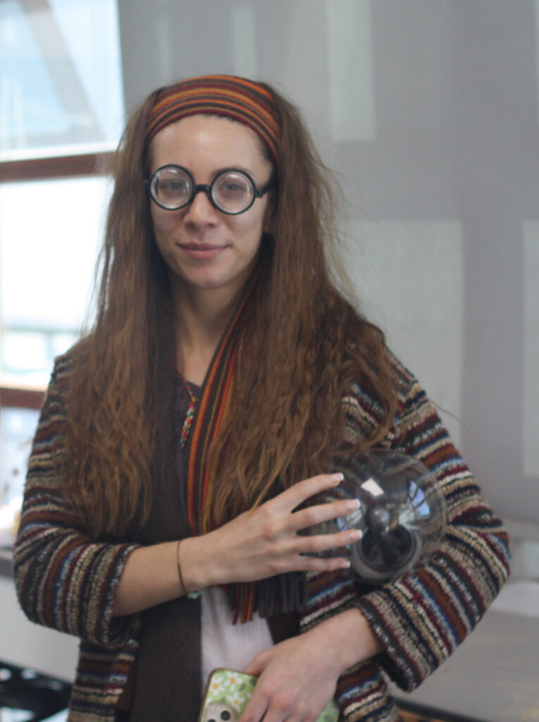 A person wearing thick round glasses, a colorful striped headband, and a patterned cardigan, holding a clear glass orb.